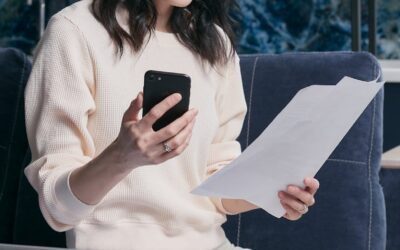 Une jeune femme utilise son smartphone et son ordinateur portable pour gérer ses finances, semblant concentrée et préoccupée.