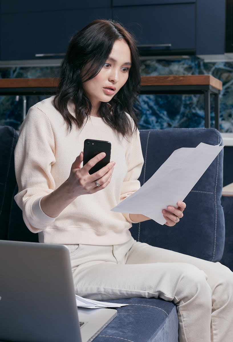 Une jeune femme utilise son smartphone et son ordinateur portable pour gérer ses finances, semblant concentrée et préoccupée.