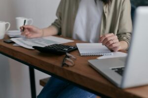 Une femme gère ses finances à la maison, à l'aide d'un ordinateur portable et d'une calculatrice sur un bureau en bois.