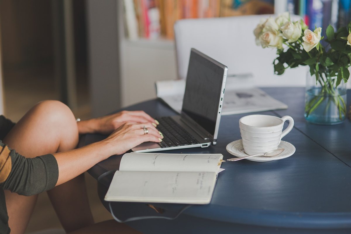 Une personne tape sur un clavier d'ordinateur portable à côté d'un carnet et d'une tasse de café blanche